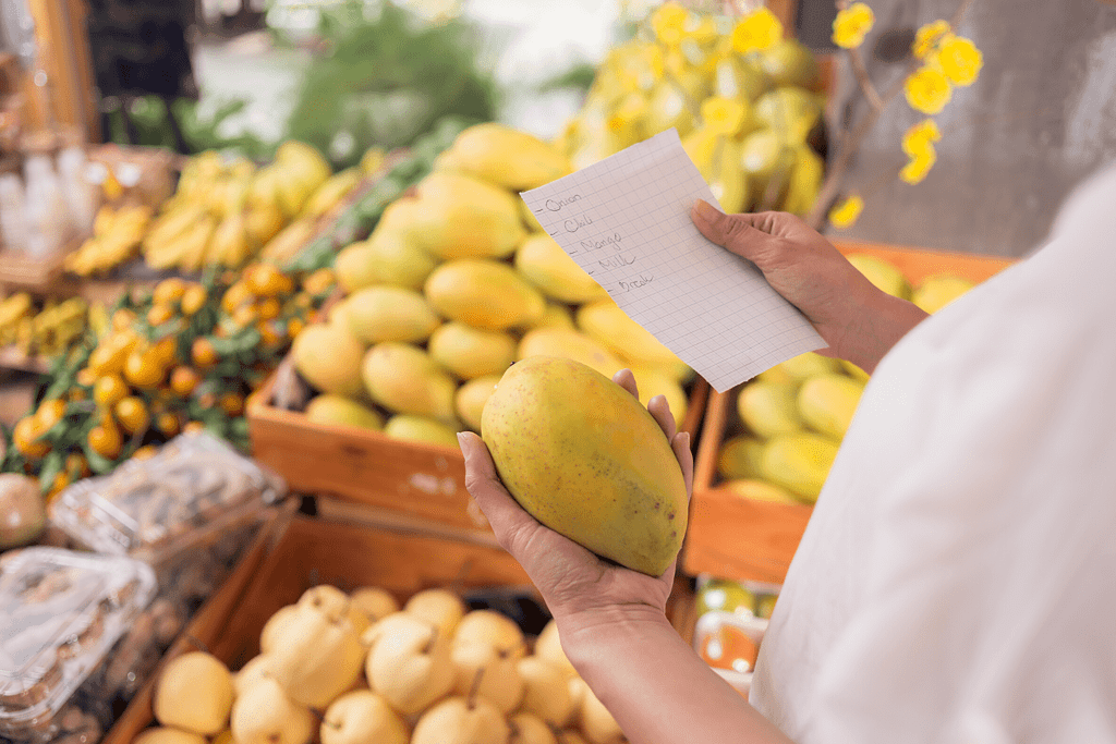 person in grocery store holding mango in one hand and shopping list in the other hand 
